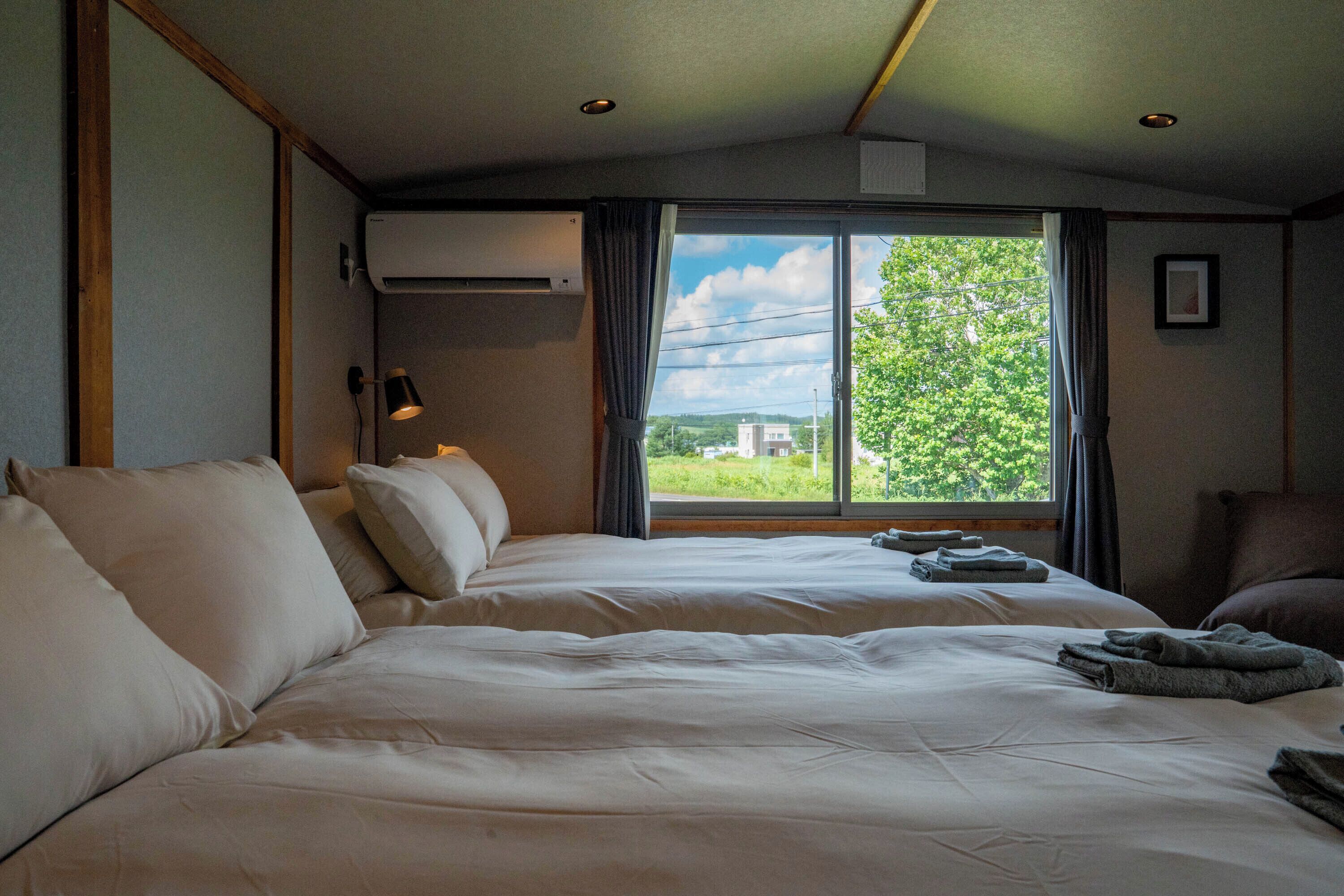 A hotel room with two beds, a window with curtains, an air conditioning unit, and a lamp.