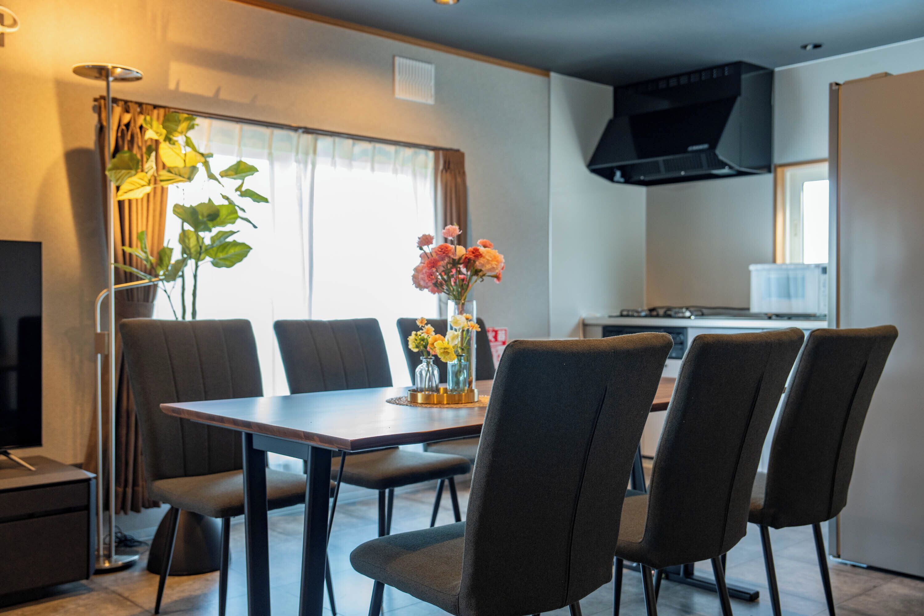 A dining area with a table and chairs, a vase with flowers, and a kitchen in the background.