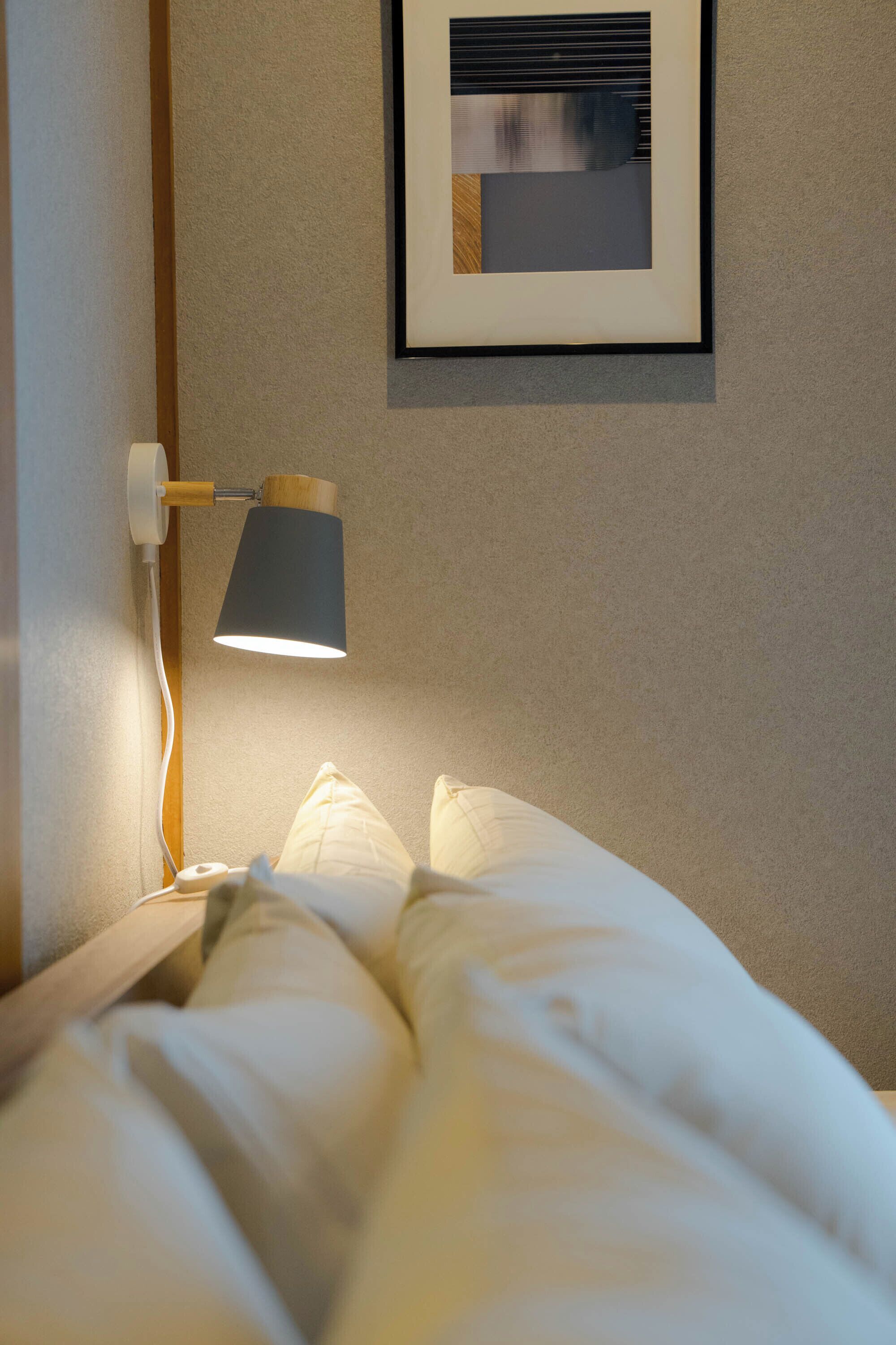 Un lit bien fait, avec du linge de lit blanc, une lampe fixée au mur et un tableau encadré accroché au mur.
