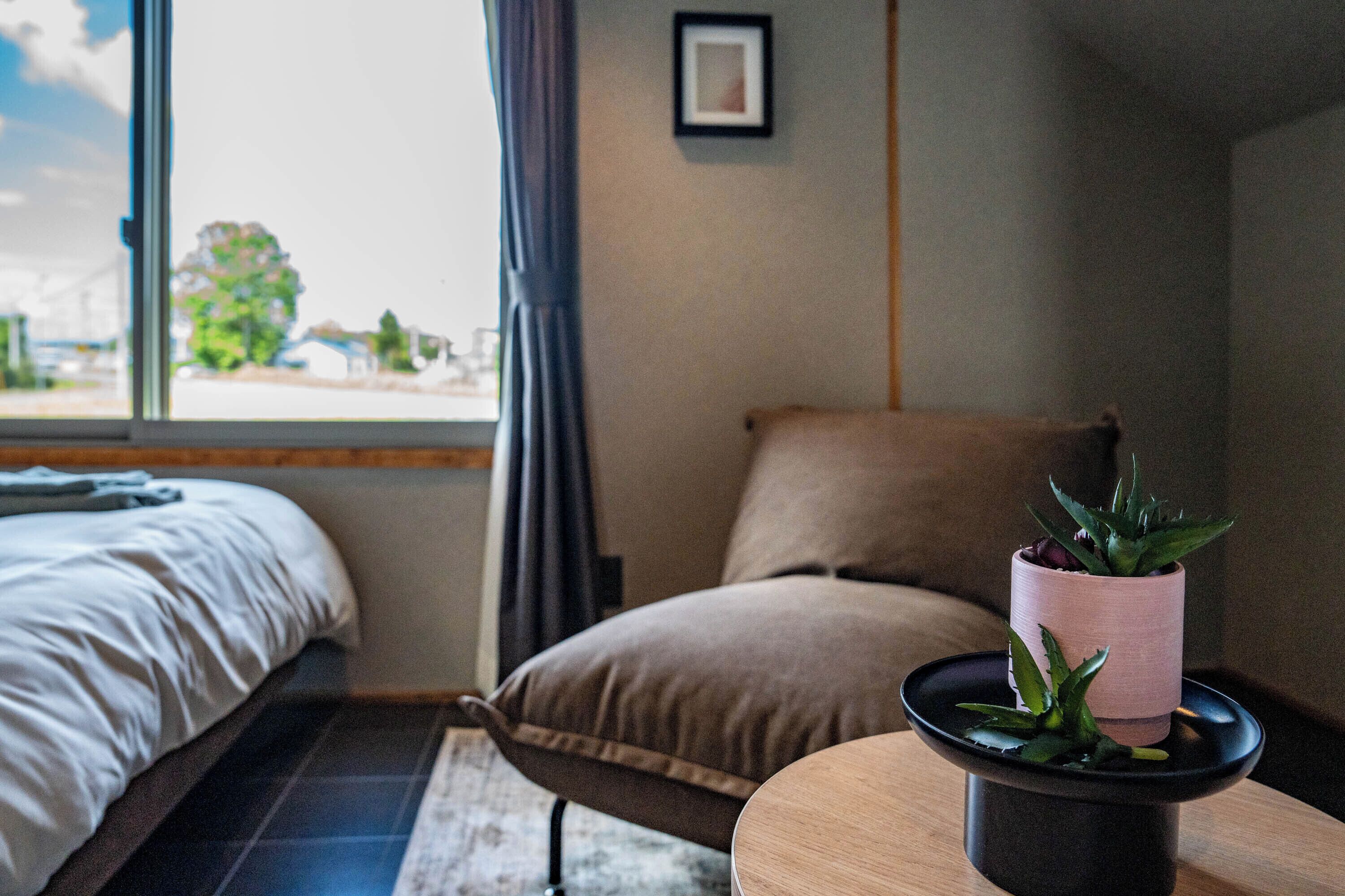 A bedroom with a bed, a chair, a small table with a plant, and a window with a view of buildings and trees.