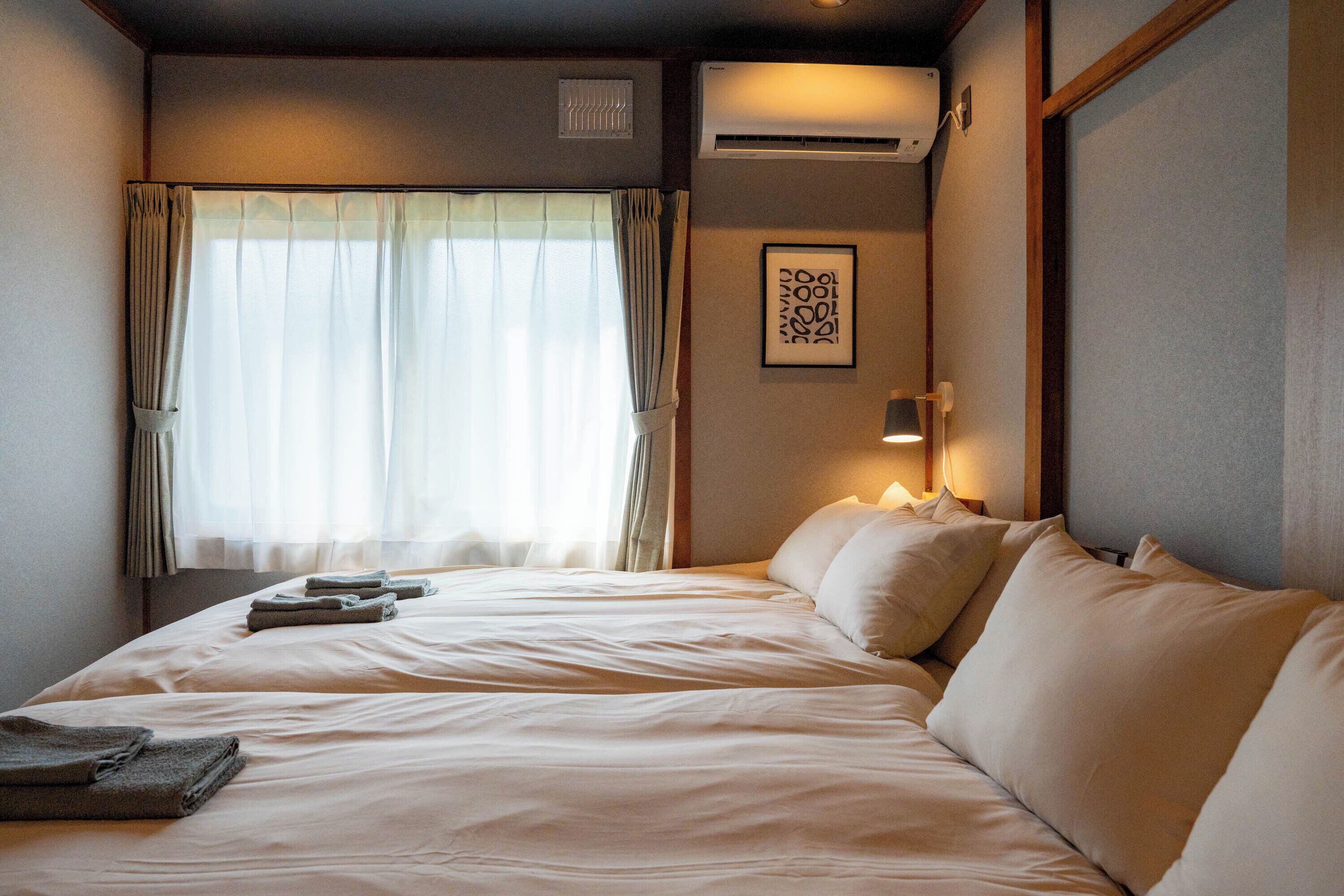 A hotel room with a large bed, a bedside table, a wall-mounted air conditioner, a window with curtains, and a framed picture on the wall.