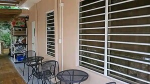 A covered outdoor area with a table and chairs, a shelving unit with items, and large windows with horizontal blinds.