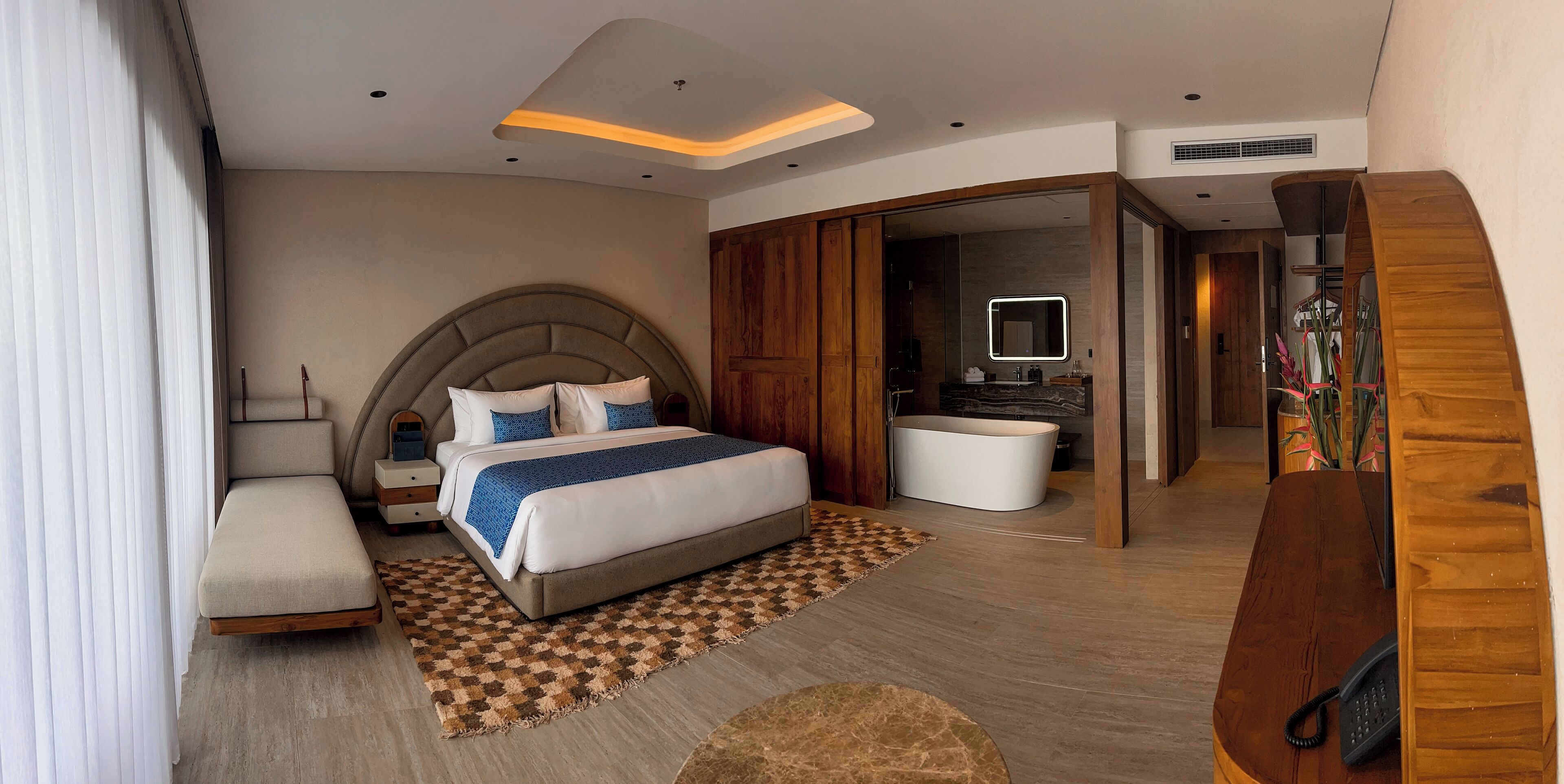 A modern hotel room with a large bed, a separate sitting area, a bathroom with a mirror, and a wooden staircase.