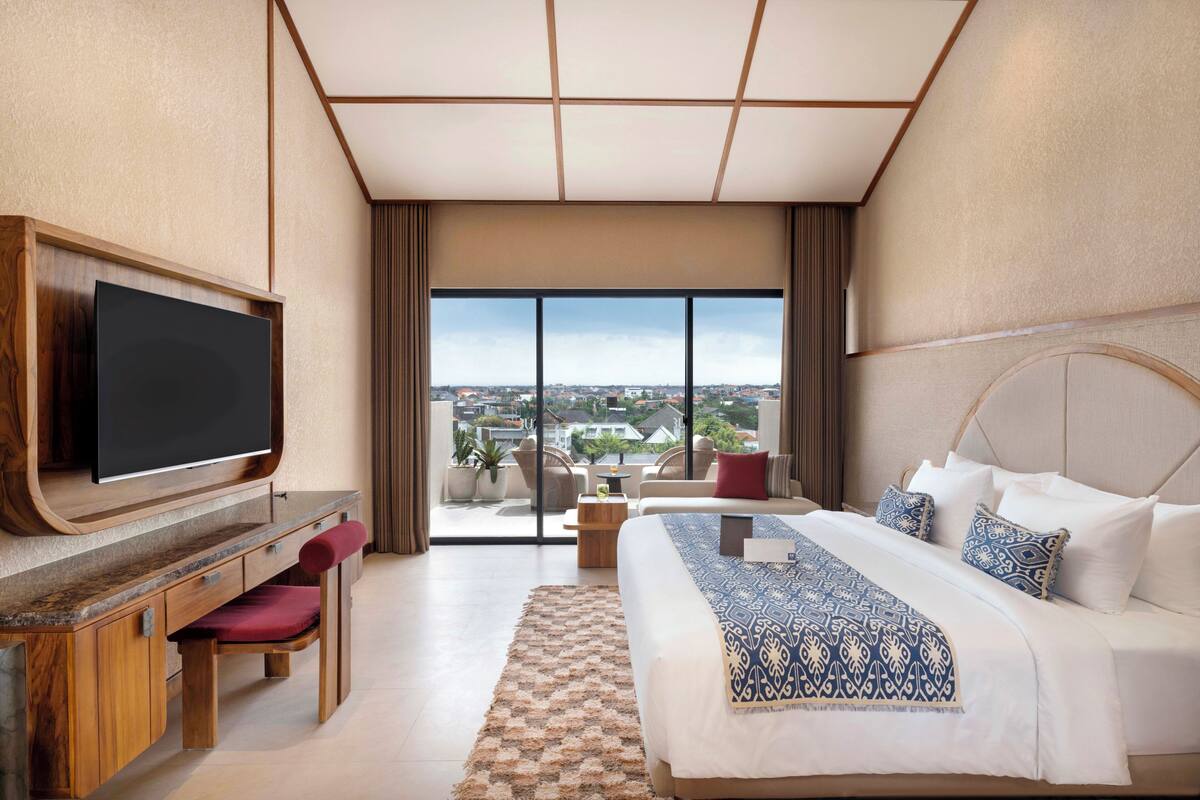Une chambre d’hôtel avec un grand lit, un téléviseur fixé sur un meuble en bois, un coin salon avec un fauteuil rouge et une vue sur la ville grâce à de grandes fenêtres.