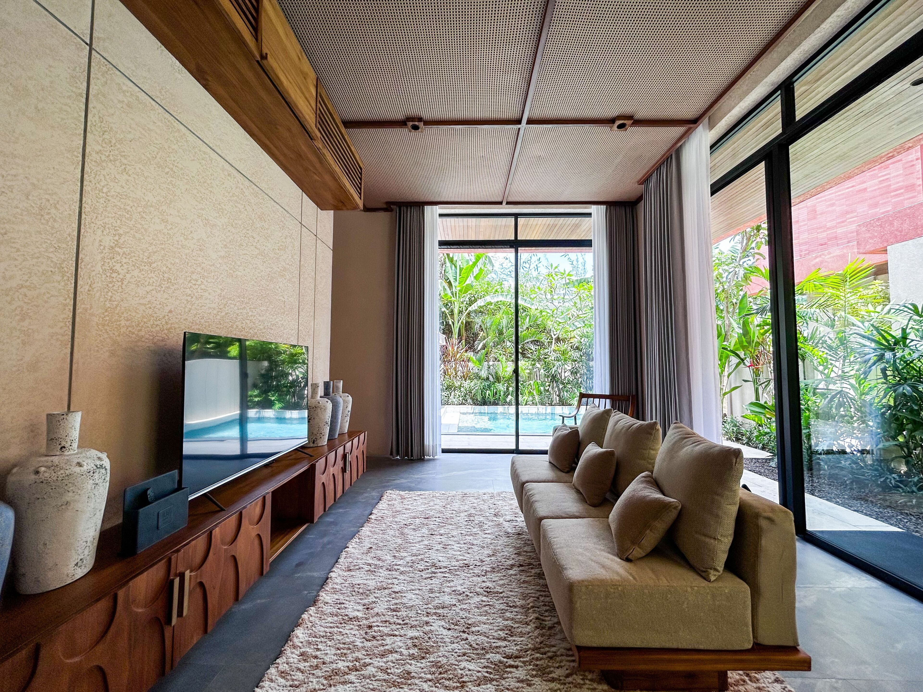 A modern living room with a large flat-screen TV, a wooden console table, a beige sofa, and a view of a swimming pool and greenery.