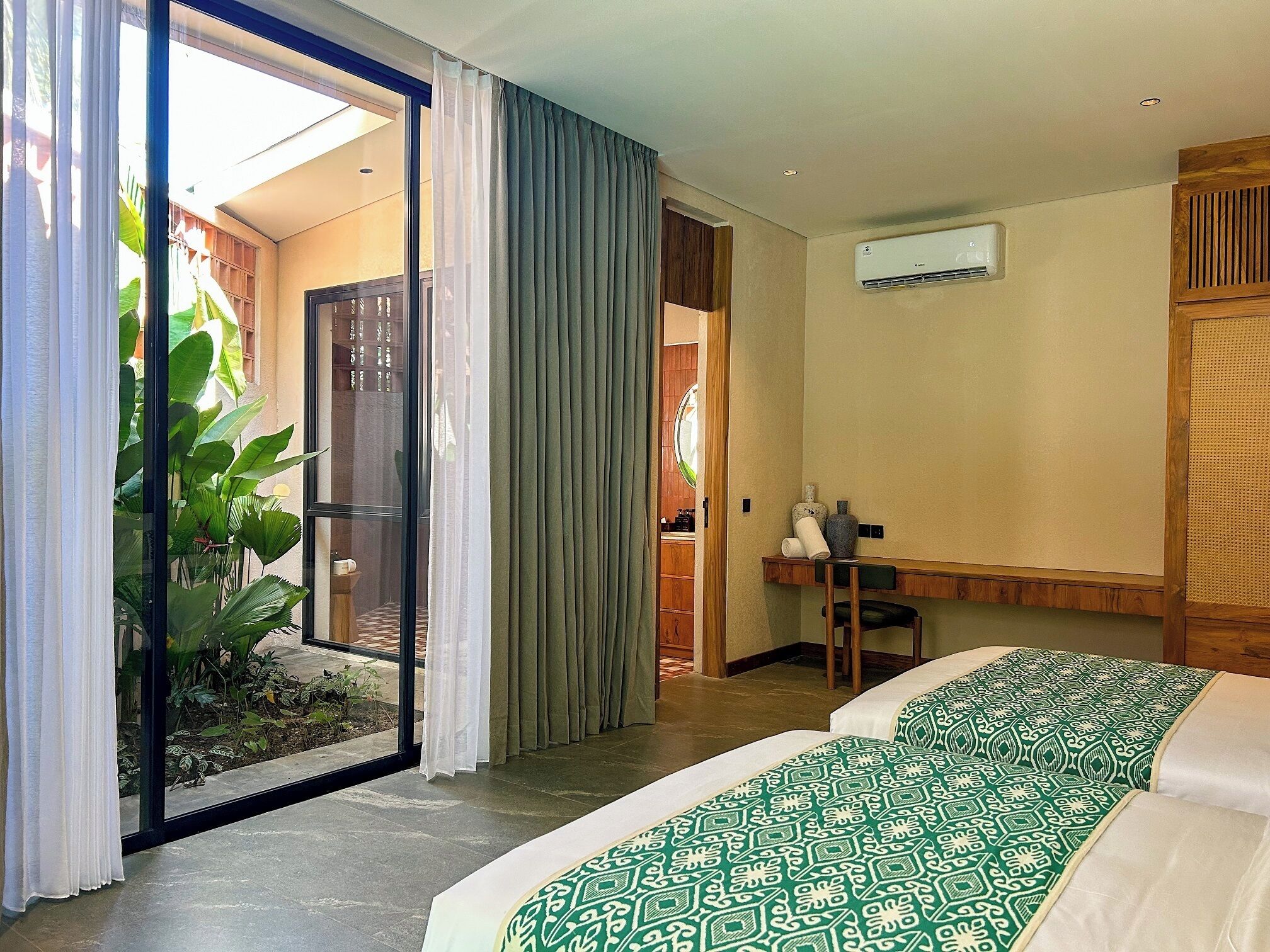 Une chambre d’hôtel équipée d’un lit, d’un bureau, d’un climatiseur et offrant une vue sur un jardin grâce à une porte vitrée.