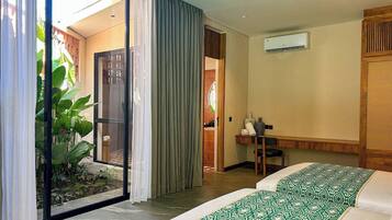 Une chambre d’hôtel équipée d’un lit, d’un bureau, d’un climatiseur et offrant une vue sur un jardin grâce à une porte vitrée.