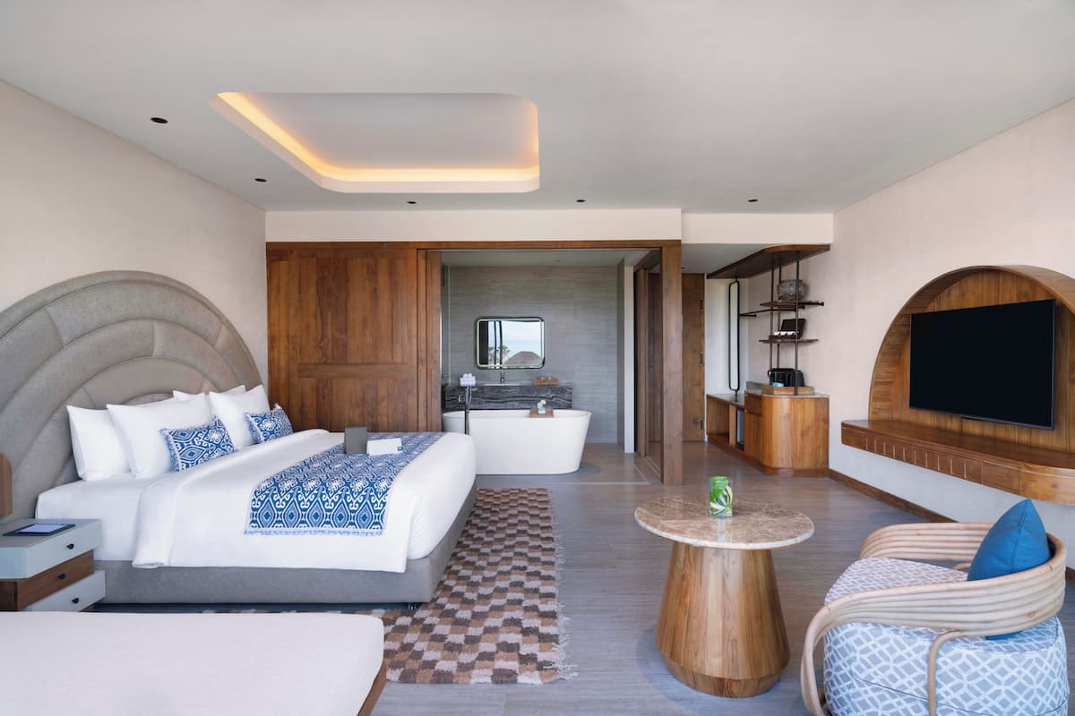 Une chambre d’hôtel moderne dotée d’un grand lit, d’un coin salon, d’une télévision et d’une salle de bain visible à travers une porte ouverte.