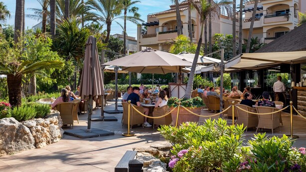 An outdoor dining area with wicker furniture and umbrellas, surrounded by palm trees and a building with balconies.