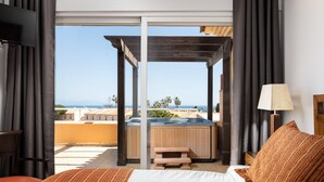 A hotel room with a bed, a wooden bench, a hot tub, and a view of the sea.