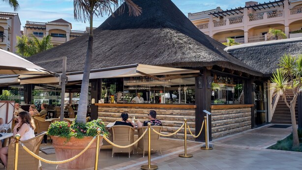 An outdoor dining area with a thatched-roof structure, wicker furniture, and a bar.