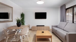 A modern living room with a round wooden table, beige chairs, a grey sofa, and a flat-screen TV mounted on the wall.