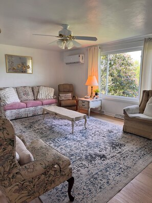 Un salon avec un ventilateur de plafond, un canapé, un fauteuil, une table basse et une fenêtre avec des rideaux.