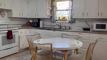 A kitchen with white cabinets, a round dining table with chairs, a ceiling fan, and a window with a curtain.