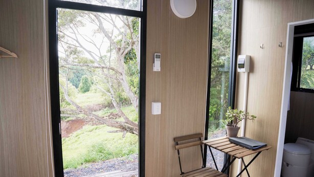 Quarto com porta de vidro deslizante para um jardim, uma pequena mesa com um laptop, uma cadeira e um vaso de planta.