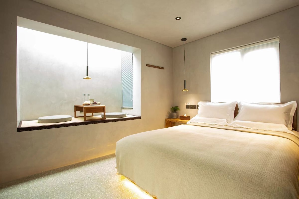 Une chambre moderne avec une grande fenêtre, une table de chevet avec une lampe et un lit recouvert de linge de lit blanc.