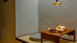 Ein schlichter Raum mit Tatami-Boden, einem niedrigen Holztisch mit Obstschale, einem Teekessel und einem kleinen Hocker.