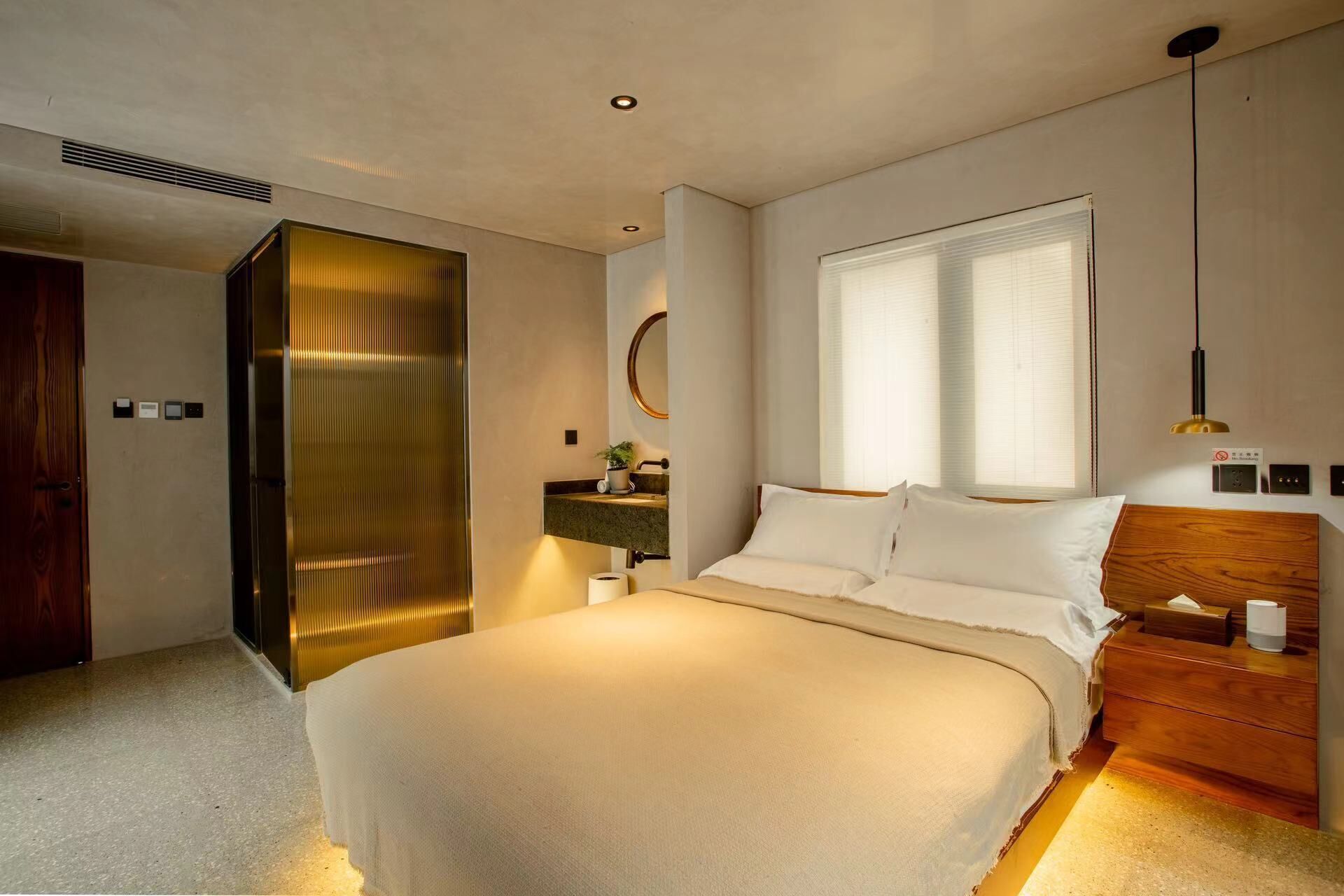 A modern bedroom with a large bed, a bedside table, a nightstand, a window with blinds, and a wall-mounted mirror.