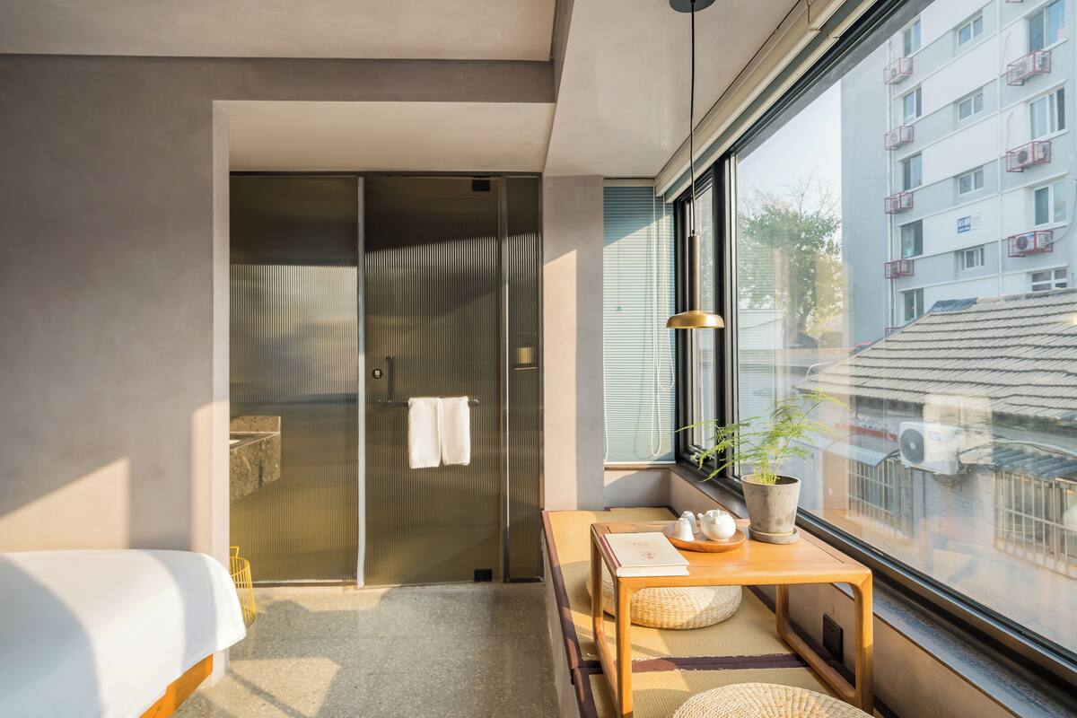 Une chambre d’hôtel moderne dotée d’un placard avec une porte coulissante en verre, d’une table de chevet en bois agrémentée d’une plante, et offrant une vue sur la ville par la fenêtre.