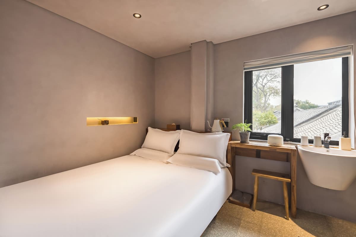 Une chambre moderne avec un lit, une table de chevet et une fenêtre donnant sur des immeubles.