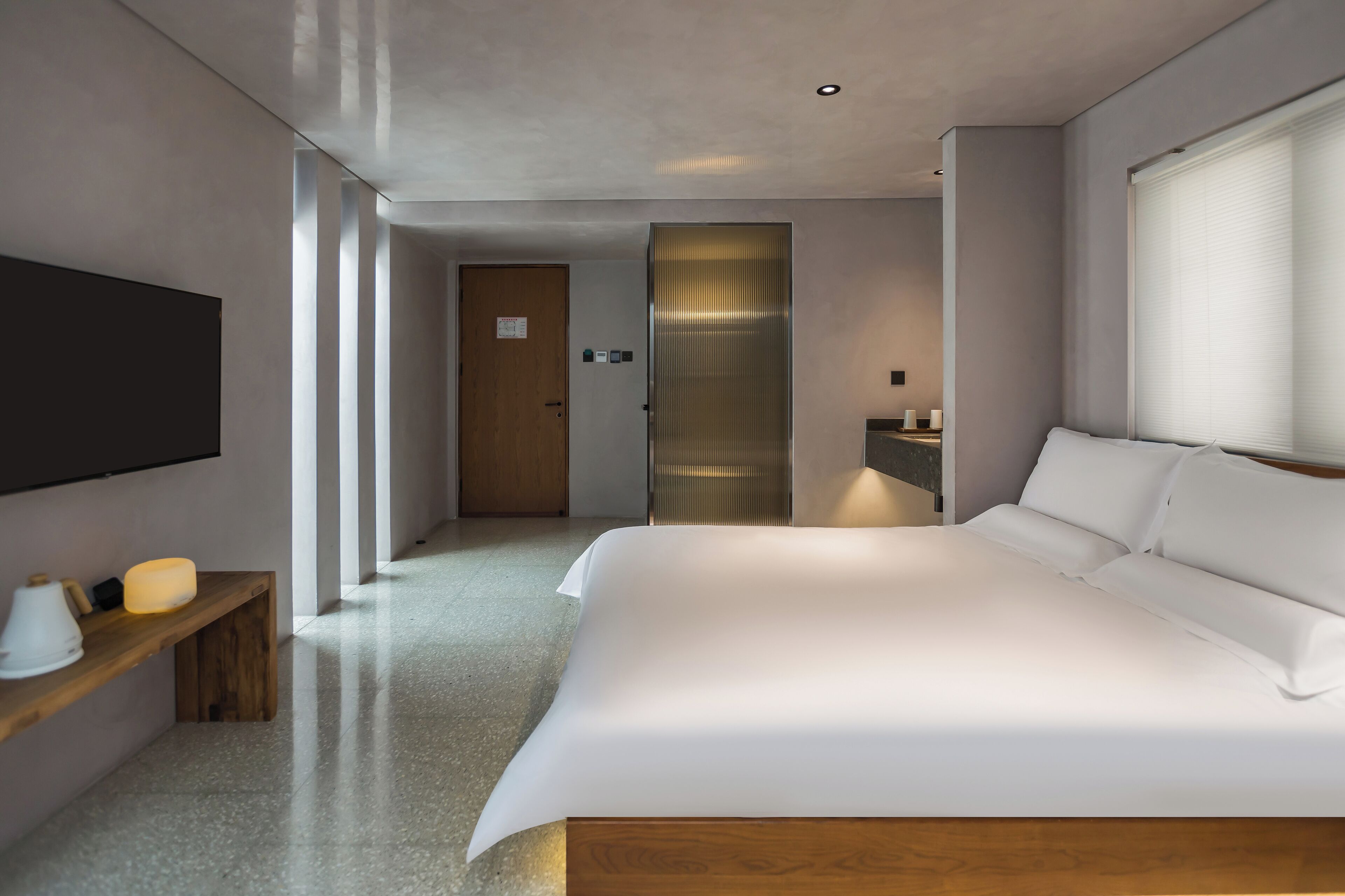 A modern hotel room with a large bed, a flat-screen TV mounted on the wall, a bedside table with a lamp, and a wooden bench.