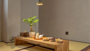 Ein Raum mit Tatami-Matten, einer niedrigen Holzbank, einem Blumentopf, einem Teeservice und einem kleinen Tisch mit Snacks.
