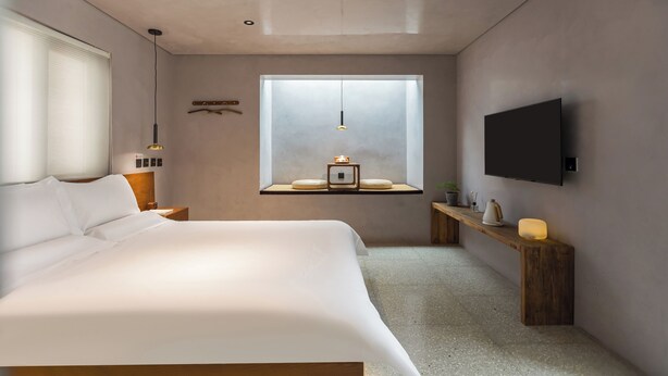 Ein modernes Schlafzimmer mit einem großen Bett, einem Nachttisch, einem kleinen Fenster mit Aussicht, einem angebrachten Fernseher und einer Deckenleuchte.