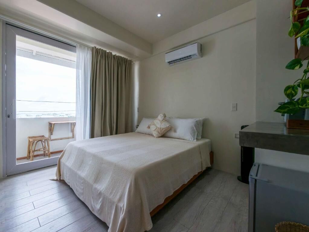 Quarto com cama, janela com cortinas, ar condicionado e uma pequena mesa com planta.