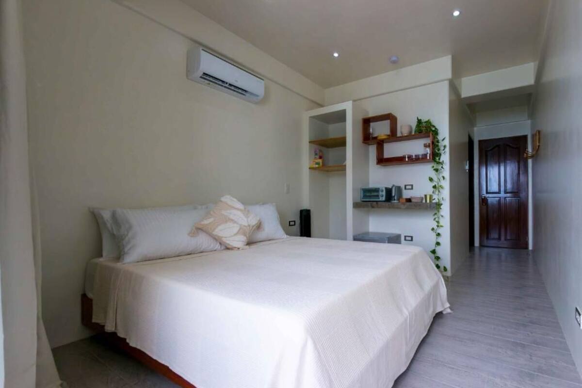 A bedroom with a bed, an air conditioning unit, a bedside table, and a door leading to another room.