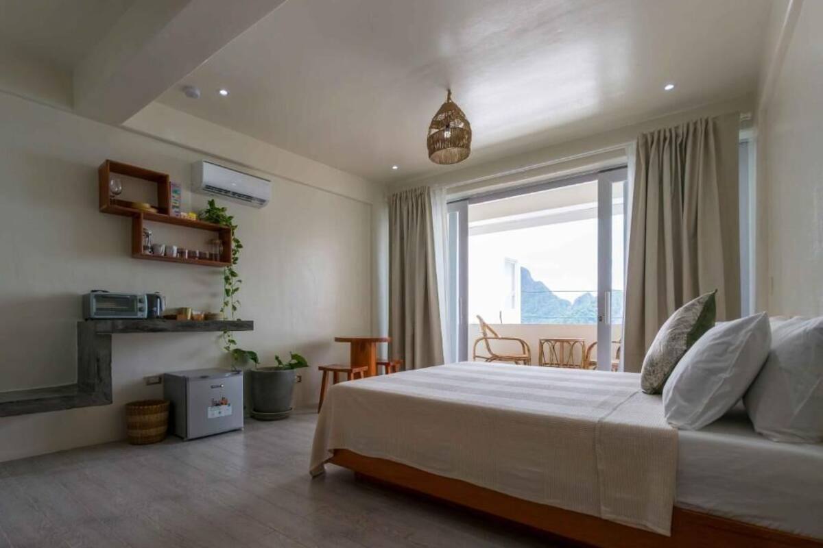A bedroom with a bed, a small table, a chair, a potted plant, and a view of the outdoors.