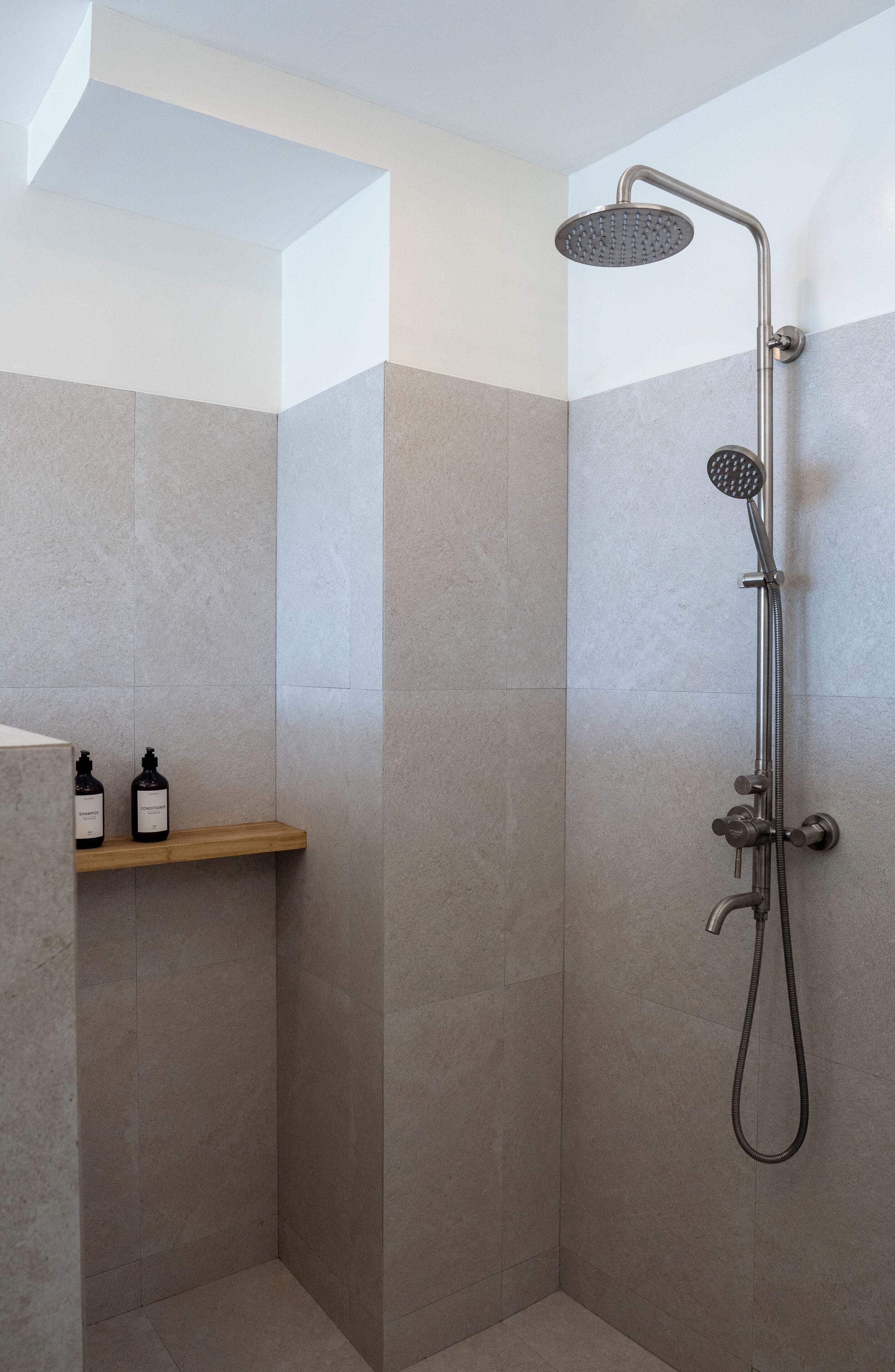 Een douche met een handdouche en regendouche, een houten plank met twee flessen, en een hoek met een wit plafond.