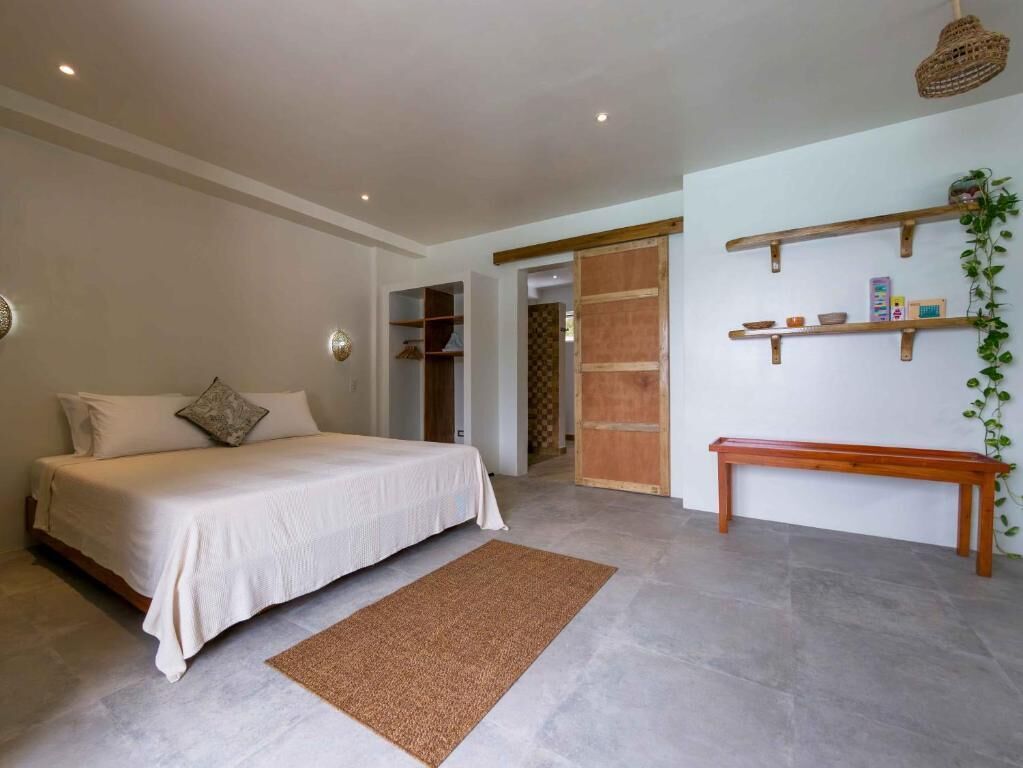 Une chambre à coucher comprenant un lit, un banc en bois, une étagère fixée au mur et une porte donnant sur une autre pièce.