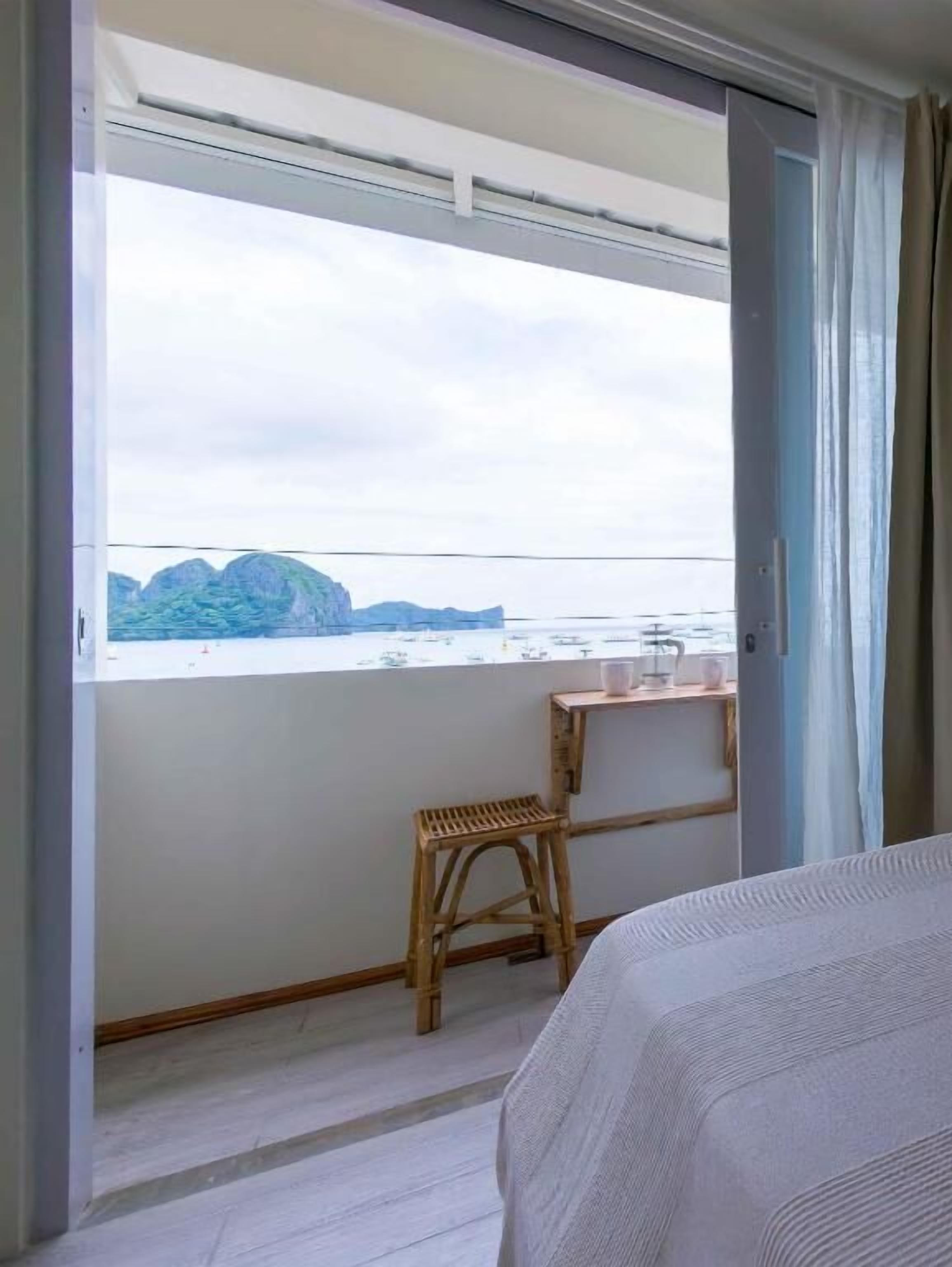 Una habitación con una cama, un taburete de madera, una pequeña mesa con una taza y vistas a un cuerpo de agua con montañas al fondo.