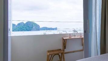 Una habitación con una cama, un taburete de madera, una pequeña mesa con una taza y vistas a un cuerpo de agua con montañas al fondo.