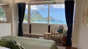 Quarto com uma cama, uma cadeira, uma mesinha e vista para o oceano e montanhas.