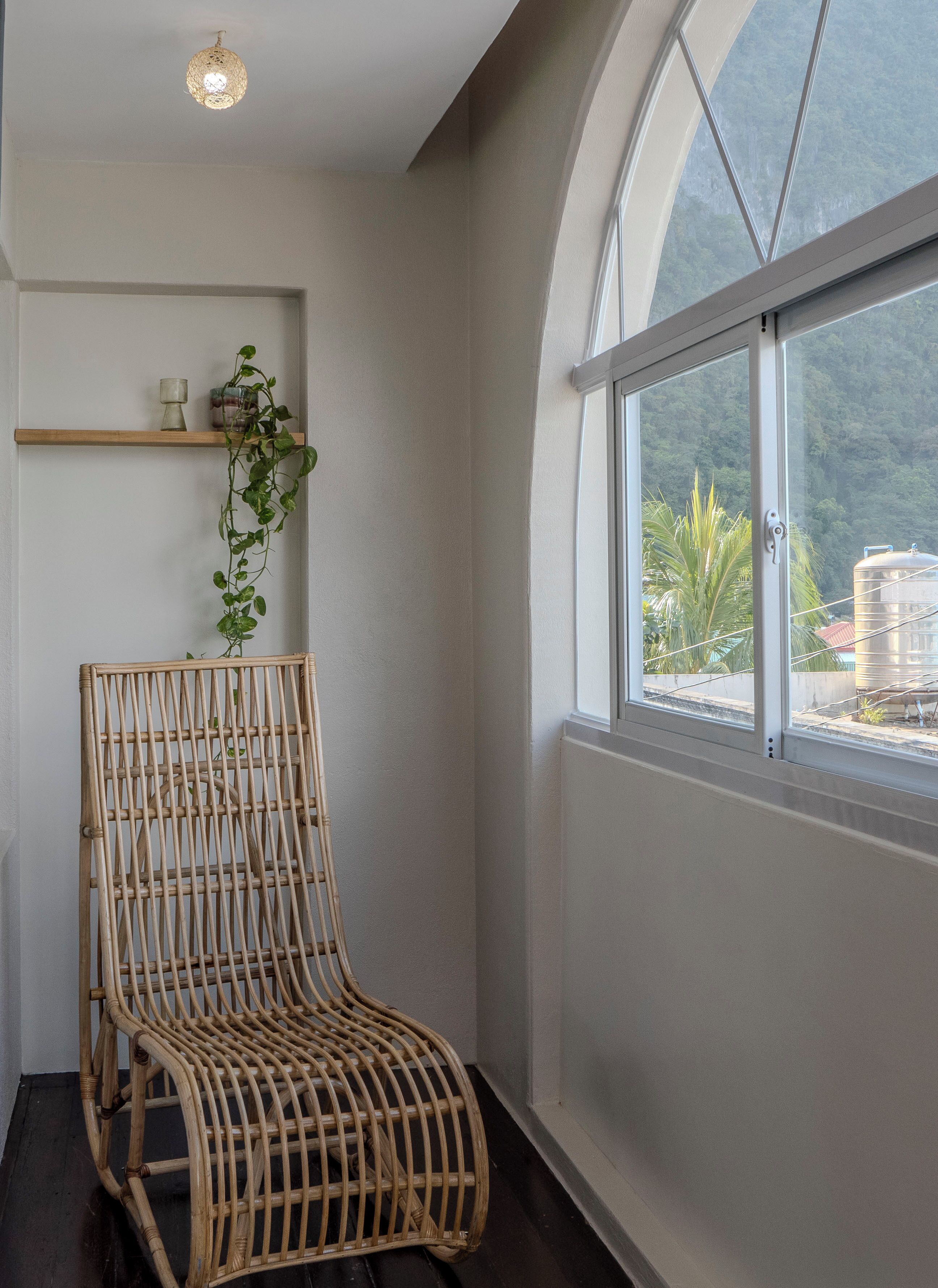 A room with a wicker chair, a small shelf with decorative items, and a large window offering a view of greenery and a water tank outside.