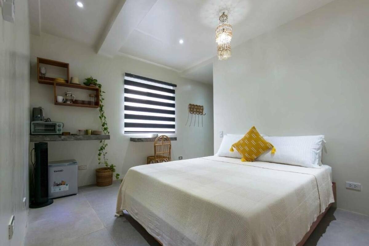 A bedroom with a bed, a small kitchenette, a window with blinds, and a chandelier.