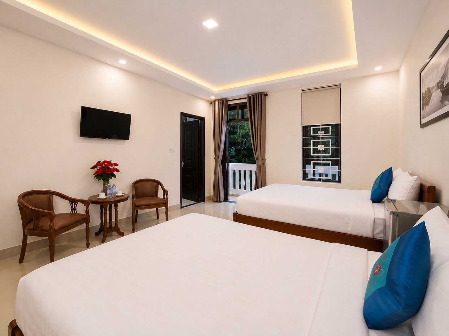 A hotel room with two beds, a TV, a small table with a vase of flowers, and a view of the outdoors.