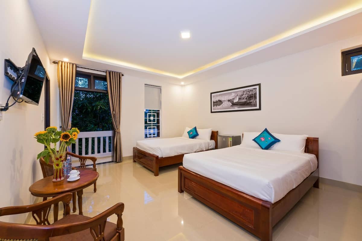 A hotel room with two wooden beds, a TV mounted on the wall, a dining table with chairs, and a balcony with curtains.