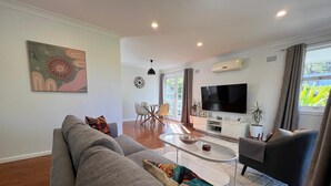 A modern living room with a grey sofa, a coffee table, and a TV. There is a dining area with chairs and a table, and a plant on the windowsill.