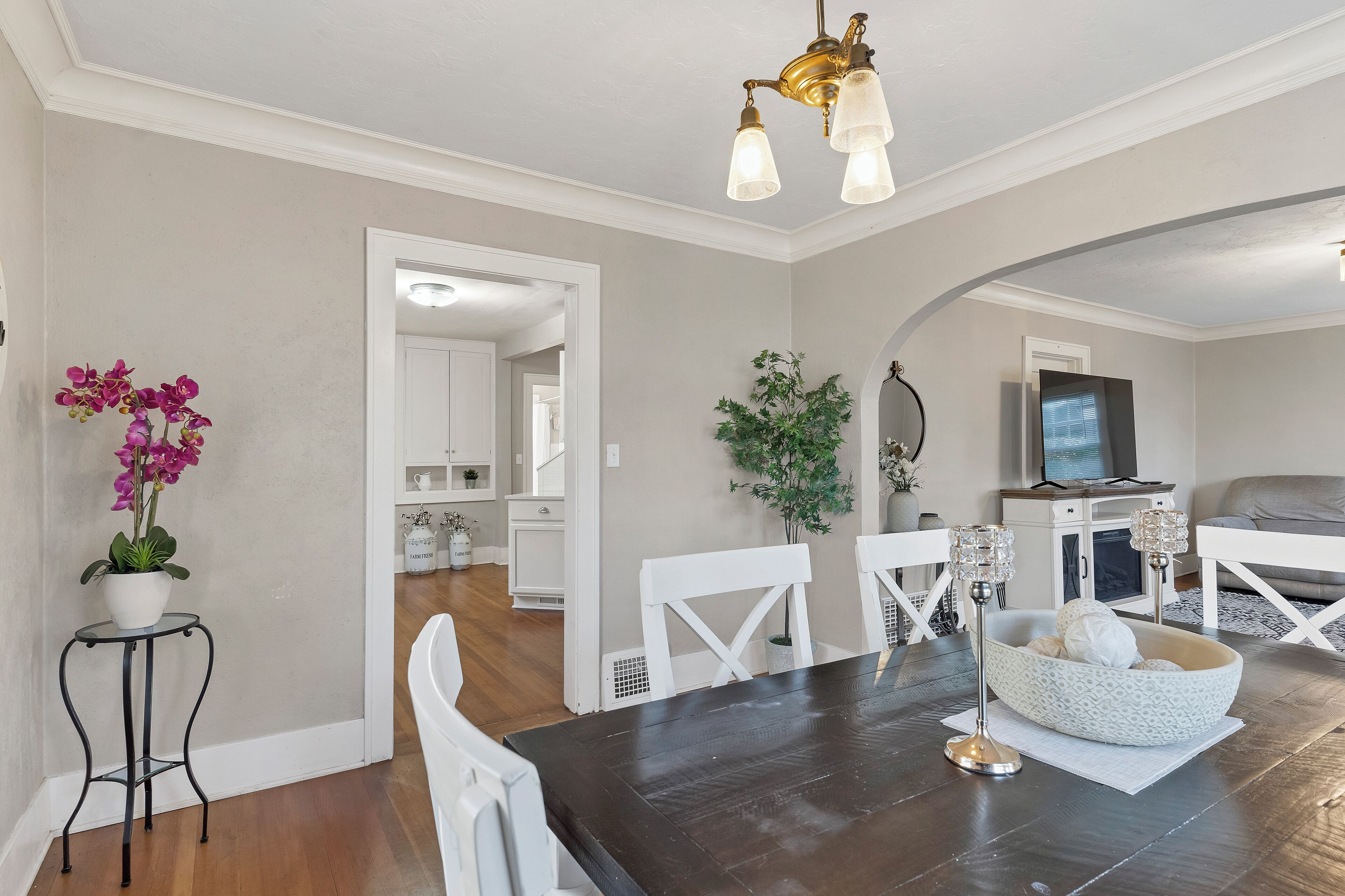 A dining area with a dark wooden table, white chairs, and a white patterned centerpiece. There is a potted orchid on a small table to the left.