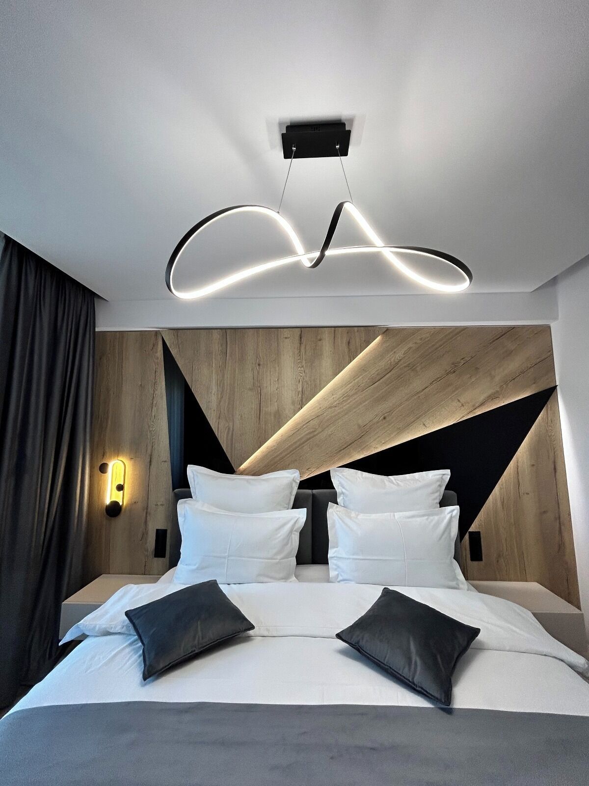 A modern bedroom with a bed, white bedding, and black pillows. There is a wall-mounted light fixture above the bed and a small bedside table with a lamp.
