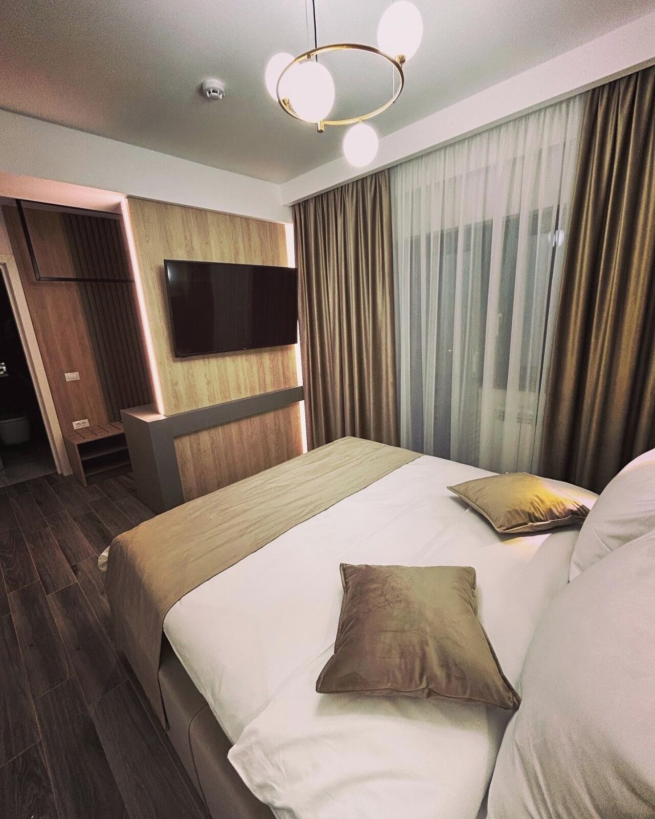 A modern hotel room with a large bed, a flat-screen TV mounted on the wall, and a separate seating area with a wooden panel design.