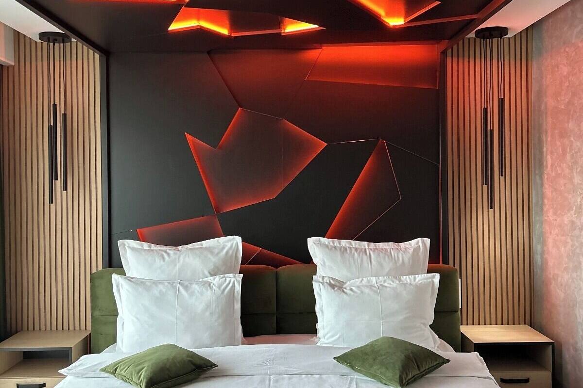 A modern bedroom with a geometric ceiling design, a large bed with white and green bedding, and wooden paneling on the walls.