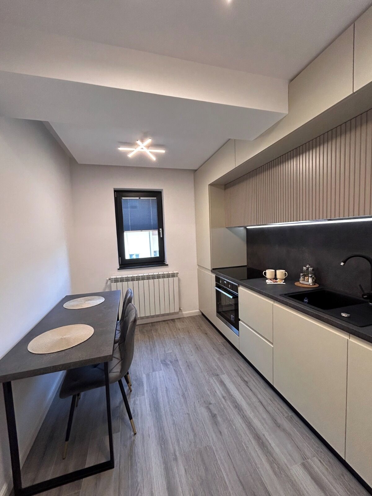A compact kitchen with a dining area, featuring a modern stove, a black countertop, and a window with blinds.