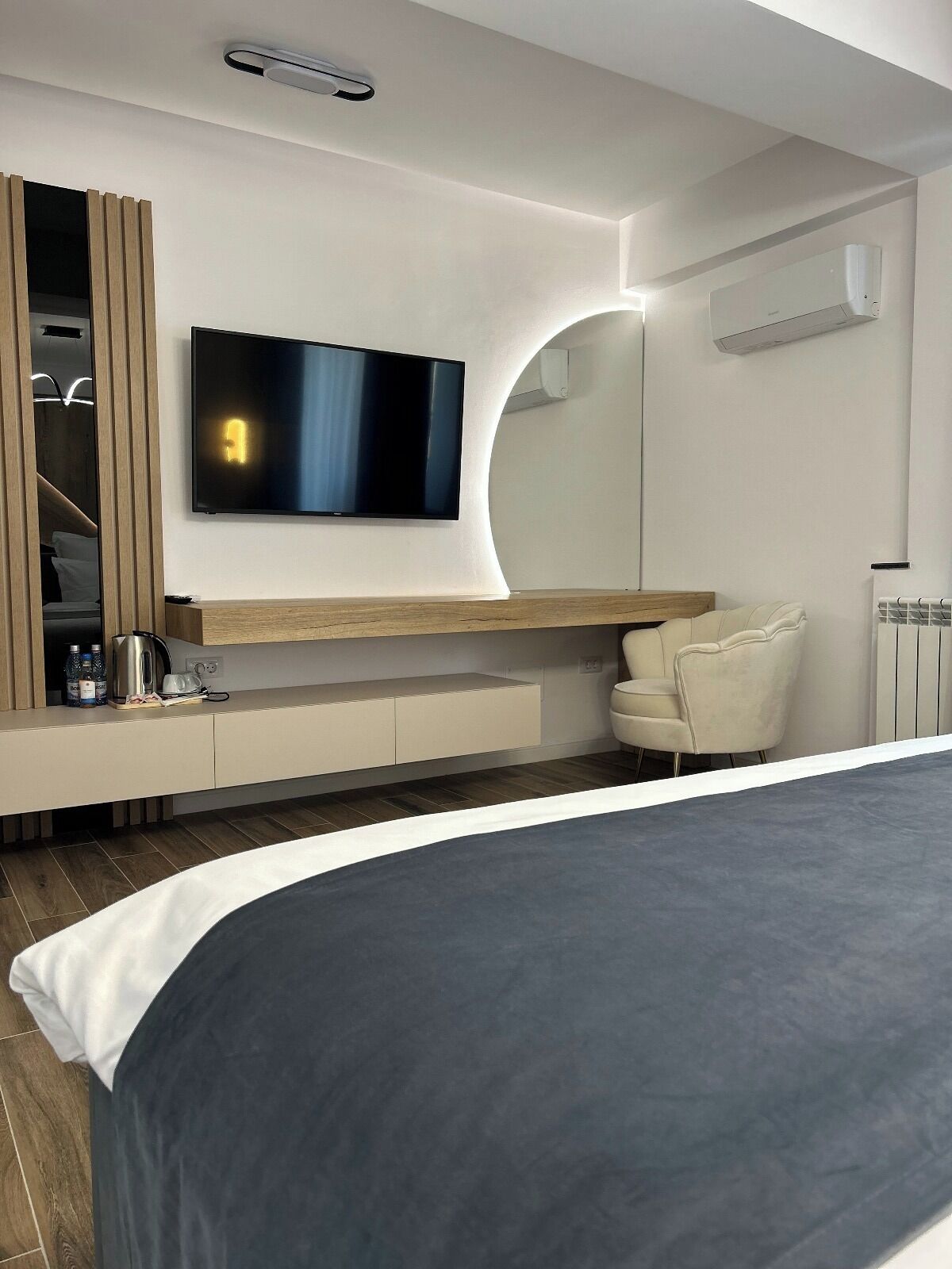 A modern bedroom with a large bed, a flat-screen TV mounted on the wall, a built-in wardrobe, and a seating area with a chair.