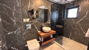 Un baño moderno con pared de mármol, espejo redondo, tocador de madera, inodoro blanco y zona de ducha con mampara de vidrio.