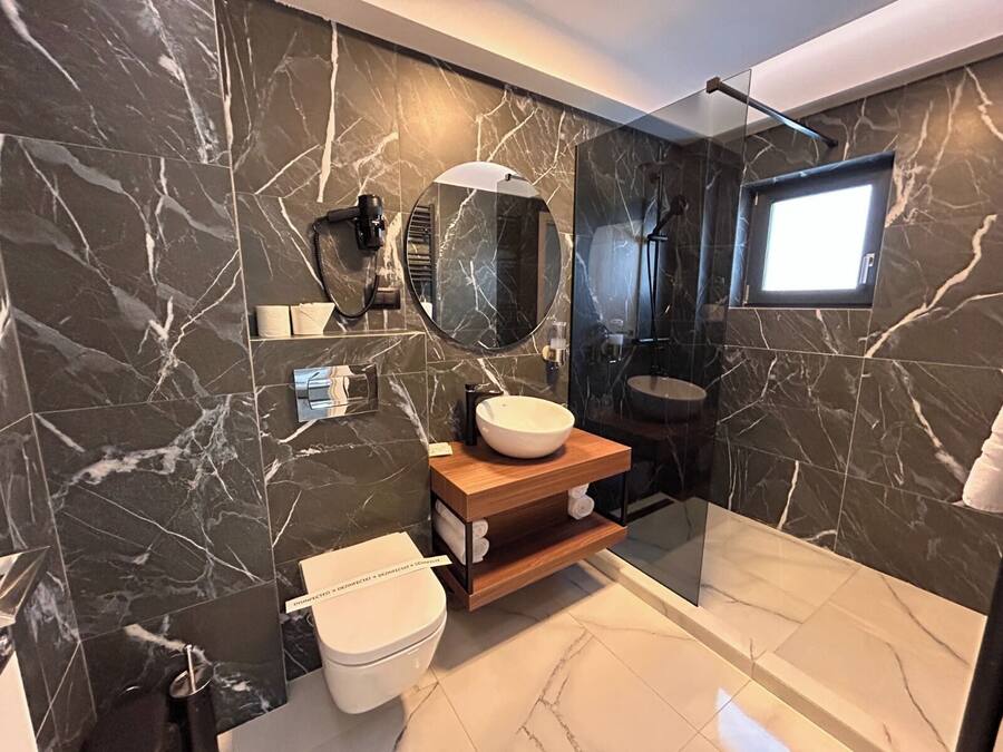 A modern bathroom with a marble wall, a round mirror, a wooden vanity, a white toilet, and a glass-enclosed shower area.