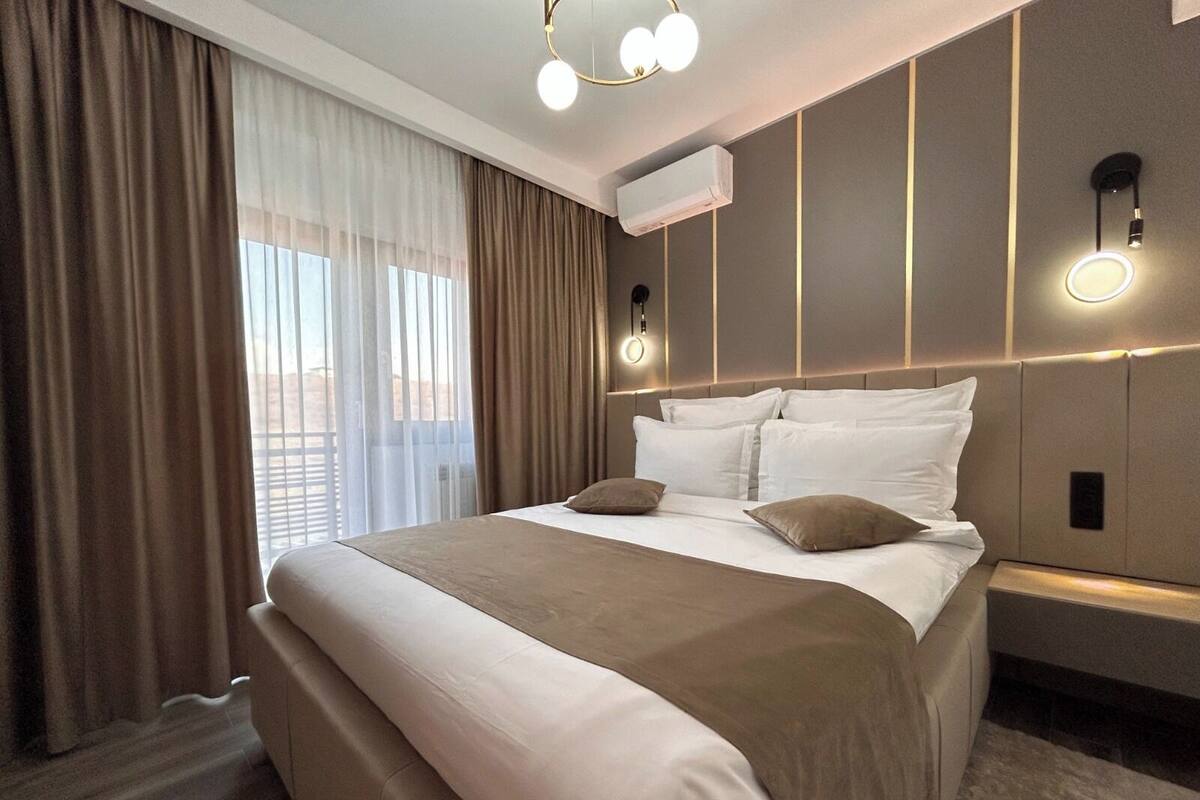 A hotel room with a large bed, a bedside table, a wall-mounted air conditioner, and a window with curtains.