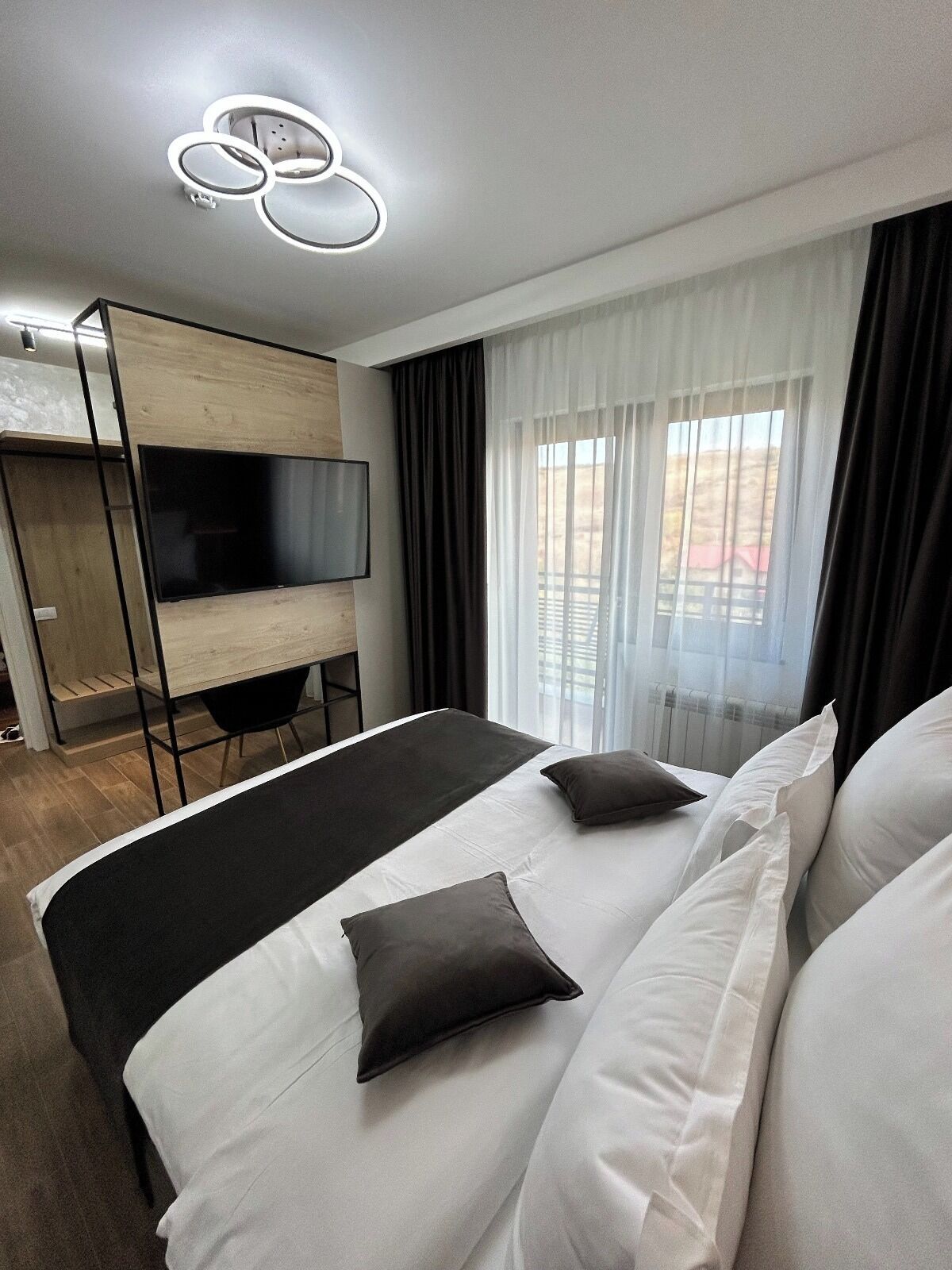 A modern hotel room with a large bed, a TV mounted on a wooden panel, and a window with sheer curtains.